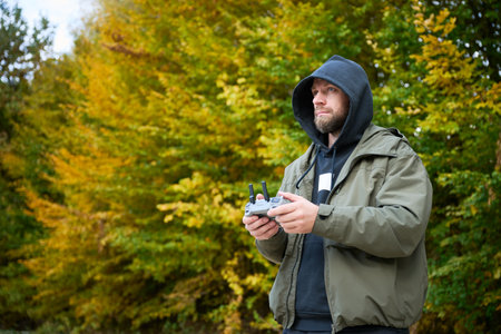 A man in a hoodie controls a drone with a remote as golden and orange leaves surround him in a...