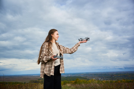 A young woman stands on a hilltop, smiling as she prepares to fly her drones. The cloudy sky adds a dramatic backdrop to her outdoor adventure.の写真素材