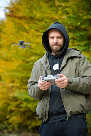 A man in a hooded jacket skillfully operates a drone in a colorful forest. The trees are ablaze with...