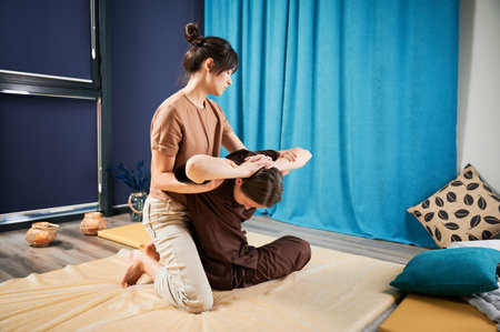 Masseuse making thai yoga massage. Woman therapist doing traditional massage treatment, stretching female patients body. Client sitting on mat, enjoying hand massage in calm atmosphere.の写真素材