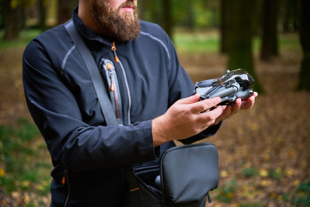 A man in a black jacket examines his drone carefully, surrounded by vibrant trees and fallen leaves...
