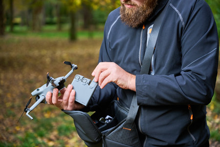 A man with a bushy beard prepares his drone for flight in a peaceful park filled with trees. He...