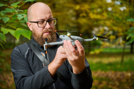 A man with a beard and glasses closely examines a drone in a vibrant park filled with colorful...