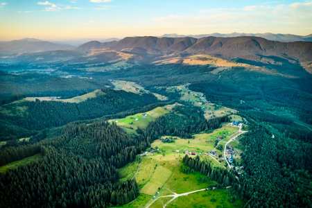 Aerial view of picturesque valley surrounded by lush green forests and rolling hills. Scattered houses and farmland are visible, creating peaceful rural scene. Sunlight casts warm glow over landscape.の写真素材