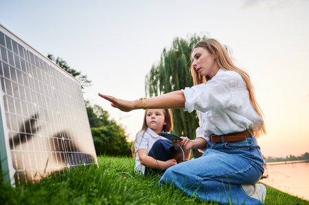 Mother connecting smartphone to photovoltaic solar panel for charging, explains its workings to daughter, sit on grass by lake. Integration of sustainable renewable energy into everyday life.の写真素材