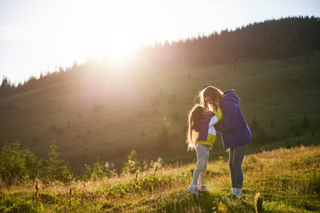 Young woman tourist spending time together with little daughter in sunlit meadow, against stunning mountain backdrop at sunset. Joyful moment captures essence of carefree fun and natural beauty.の写真素材