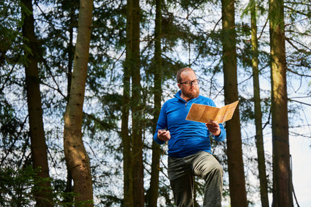 Man traveler stands in dense forest, holding map in one hand and compass in other. Tourist planning outdoor adventure or hiking route, amidst tall trees and natural surroundings.の写真素材