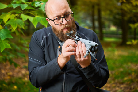 A person intently inspects a small drone while surrounded by vibrant autumn trees. Sunlight filters through the leaves, creating a peaceful atmosphere.の写真素材