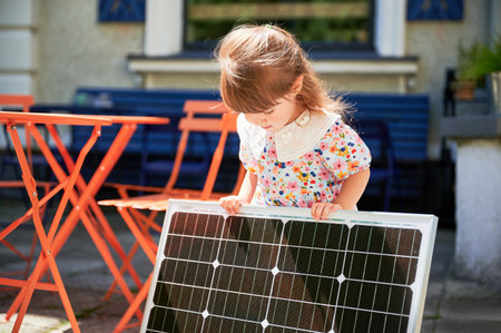 Kid holding portable solar panel for charging. Young girl in floral dress stands outside in sunny...