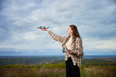 In a peaceful outdoor setting, a young woman stands on a hillside with a drone in hand, ready to take flight against a backdrop of rolling hills and a gray sky.の写真素材