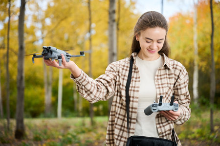 In a lively autumn forest, a woman holds her drone confidently while preparing to launch it with a...