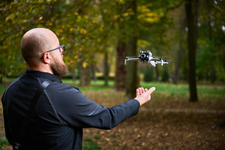 A man with a beard prepares to launch a small drone in a park surrounded by colorful autumn trees....
