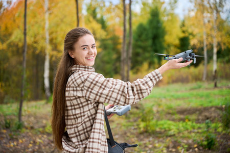 A young woman stands in an autumn forest, holding a drone in one hand and a controller in the other....