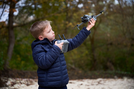 In a vibrant park setting, a young boy excitedly launches his drone into the bright blue sky while surrounded by colorful autumn leaves.の写真素材