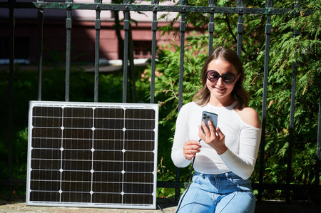 Happy woman connecting smartphone to photovoltaic solar panel. Integration of sustainable renewable energy into everyday life, demonstrating practical use of solar power for charging devices.の写真素材
