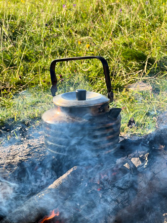 Close-up of metal kettle heating over campfire. Flames and smoke curl around well-used kettle, which...