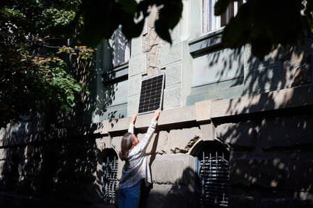 Woman holding photovoltaic solar panel in front of historical building, wearing plaid shirt and...