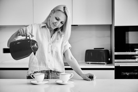 Young woman making morning coffee. Pretty female filling cup with hot water. Woman holding kettle and pouring water into cup. Black and white image.の写真素材