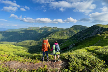 Back view of two hikers, holding hands, stand on trail overlooking mountain landscape. Tourists...
