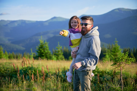 Father and his young daughter, both smiling, point towards horizon as they enjoy scenic mountain...