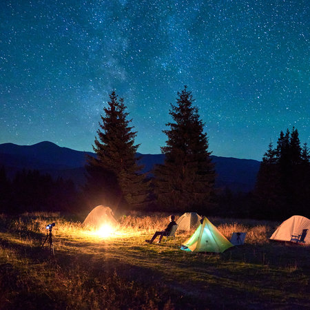 Person hiker having a rest by campfire, surrounded by glowing tents under magnificent starry sky....