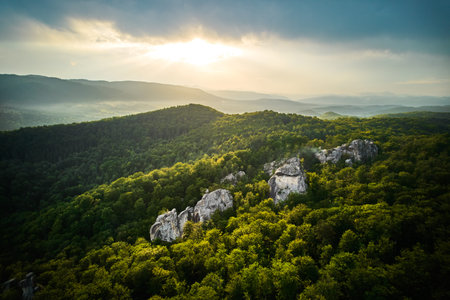 Sunlight breaks through clouds over lush, green hills, casting glow on rocky formations nestled...