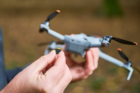 A person is holding a small memory card while standing near a drone in a wooded area. The scene...