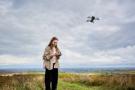 In a serene autumn landscape, a girl stands on a grassy hill, skillfully piloting a drone while...