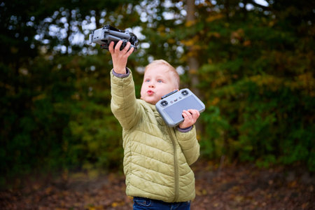 A young boy stands in an autumn forest, confidently holding a drone and controller. His excitement...