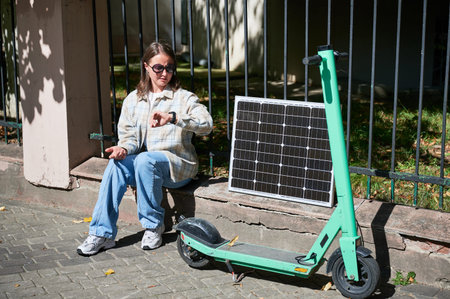 Woman checking her watch while charge electric scooter from portable solar panel, emphasizing...