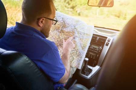 Man tourist sits in drivers seat of car, intently studying large map spread across his lap. With focused expression, traveler points to location, carefully planning his route.