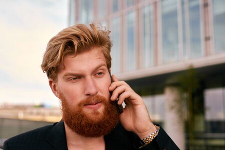 redhead talking with a grin. close up portrait hipster with red bearded. Phone conversation outdoor. Man in suit near business building.の写真素材