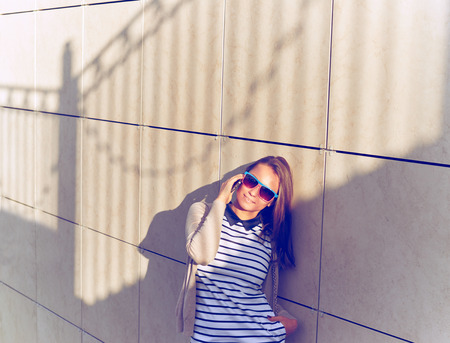 Fashionable Young Woman in Dress with stripes with her Mobile Phone While Walking a City Street. Portrait of the girl with shadows on the wall background.の写真素材