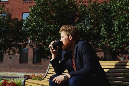 Handsome hipster drink coffee in lunch time on the bench in sunny day outdoor. Man have relax or time to think in city park.の写真素材