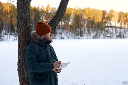 Researcher take notes in notebook outdoor in sunny winter day. Tourism concept with tourist in green jacket, red hat, who writing in book nature landscape.の写真素材