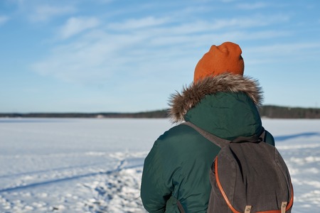 Back view on backpacker man in green winter jacket and red hat with backpack. Free space for advertising travaler goods. Free horizont with blue sky and white light clouds in sunny day.の写真素材