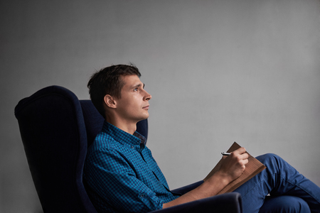 Handsome man in blue shirt and jeans sitting in dark chair and write ideas in notepad. Dreaming concept. Side view on attractive guy with grey wall background. Free space for a text or advertisment.の写真素材