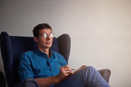 Handsome man in glasses write notes with a pen. Guy in blue shirt invent new stories. Romantic dream up a poem for darling.の写真素材