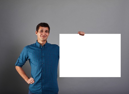 Smiling marketing manager holds big white poster in one hand. Happy man with blank paper list. Guy with copy space bord for advertising text or product.の写真素材
