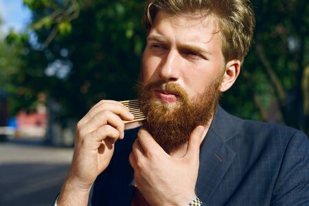 Attractive man comb hair. Hipster care his beard. Handsome caucasian boy walk outdoor in summer day. Relax time after lunch.の写真素材