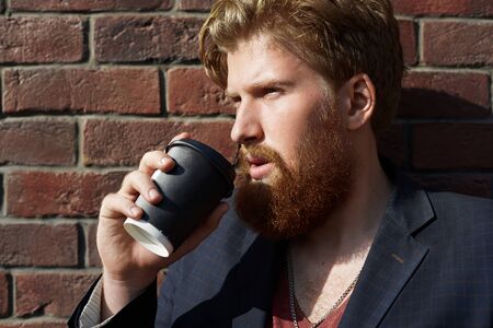 Handsome hipster drink coffee outdoor with red brick wall background. Waiting concept. Stylish dress man. Person holds cup of tea in the hand.の写真素材