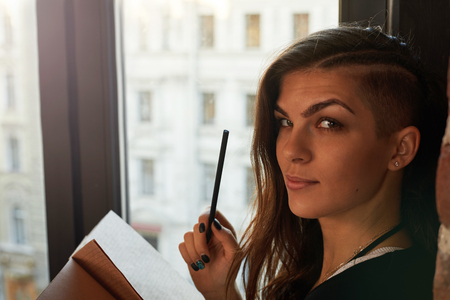 Attractive latin girl on windowsill with notebook and pencil. Woman write notes near window. Brightenning effect. Idoor view to the street.の写真素材