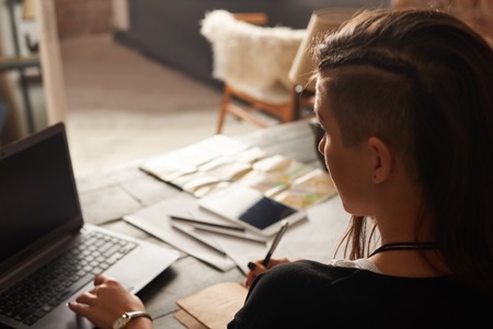 Back view on stylish latin girl, who work with laptop. Indoor loft interior. Plannin new trip with internet technologies. Tourist write notes at home.の写真素材