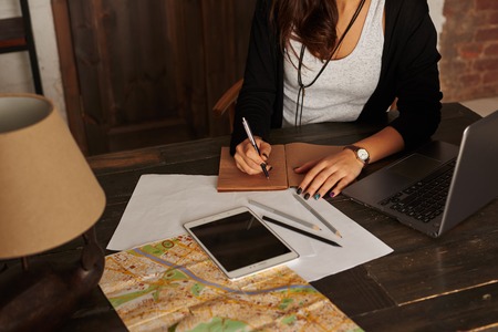 Woman planning new trip. Girl write tourist notes at home. Tablet, laptop, city map on the table. Girl learn the route in the new city. Loft interior with big wooden doors.の写真素材