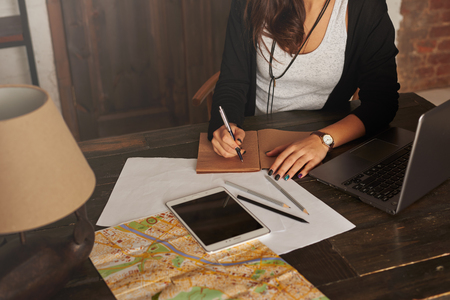 Woman planning new trip. Girl write tourist notes at home. Tablet, laptop, city map on the table. Brighten effect. Girl learn the route in the new city. Loft interior with big wooden doors.の写真素材