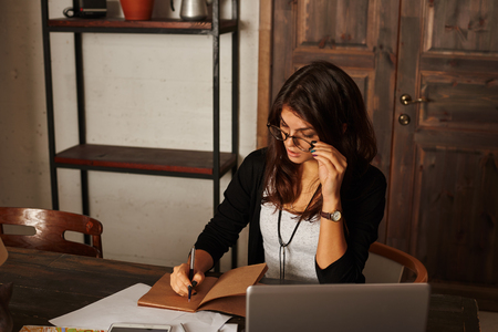 Latin pretty student work at home. Stylish beautiful woman with brunette hair in glasses write notes in notepad. Free space on the shelf for advertising products.の写真素材