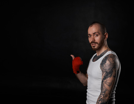 Handsome fitness man point away. Portrait of strong man with tattoo in white t-shirt. Athletic male with red bandage on kick. Kickbox concept on black background and copy space.の写真素材
