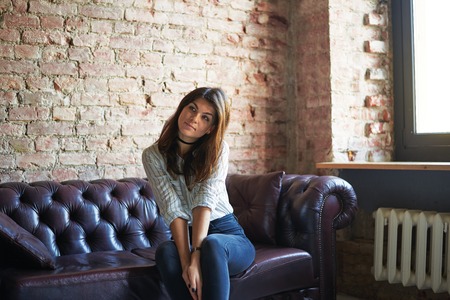 Attractive brunette sitting on couch near window at home. Portrait of woman in vintage interior witj red brick walls.の写真素材
