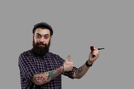 Tattooed biker show like and smoking pipe. Isolated big beard person. Attractive and smiling kind guy look into the camera. Stock worker on the break. Ready for relax time. Concept of smoke product.の写真素材