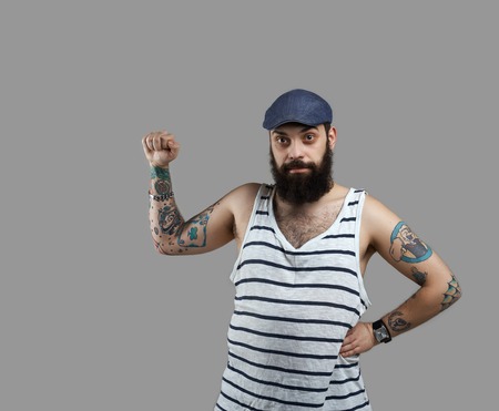 Brutal beard man with open hand for blank poster or advertisind product. Guy with tattooed hands in shirt and blue cap look to the camera. Biker sales.の写真素材